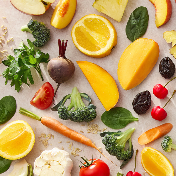 A colorful flat-lay arrangement of fresh fruits and vegetables on a light beige surface, including sliced oranges, mango pieces, broccoli florets, spinach leaves, a carrot, tomatoes, cherries, grapes, garlic, and apple slices.
