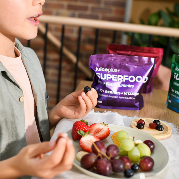 A child holds a blueberry next to a plate of fresh fruit, with two bags of juiceplus SUPERFOOD berry gummies in purple and pink packaging in the background on a table.