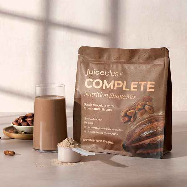 A stand-up pouch of juiceplus+ Complete Nutrition Shake Mix in Dutch chocolate flavor stands on a tabletop beside a glass of chocolate shake, a scoop of powder, and a small bowl of almonds. The scene is styled with soft window-shadow lighting against a light neutral wall.