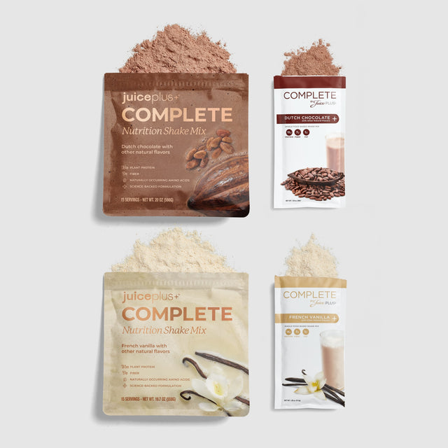 Four Juice Plus+ Complete Nutrition Shake Mix packets arranged on a light gray background, each with powder spilled above it. The packets show flavors including chocolate, vanilla, and coffee, with corresponding brown and beige powder.