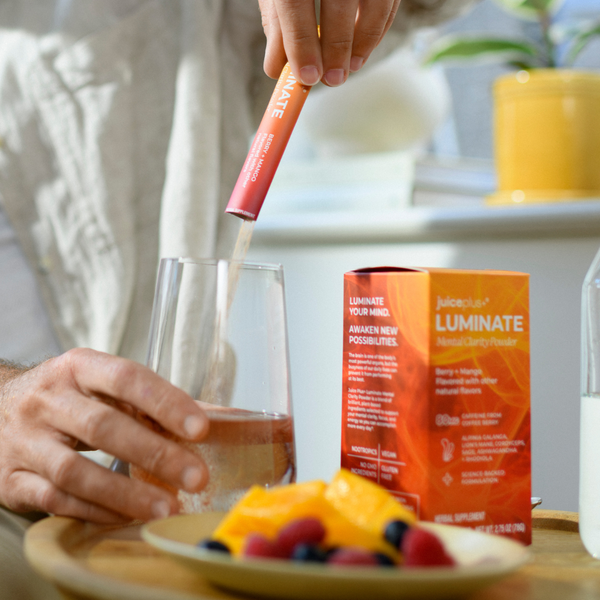 A person pours a pink liquid from a Luminate powder stick packet into a glass, with the orange Luminate box and a plate of fruit on a table in a bright indoor setting.